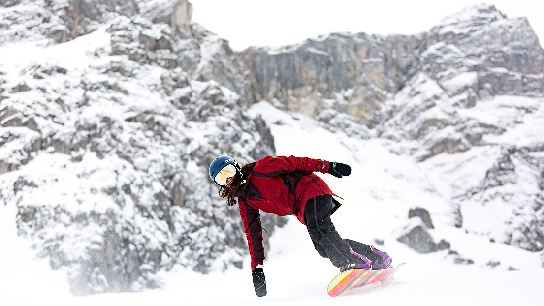 Frau mit blauem Helm und roter Jacke fährt Snowboard
